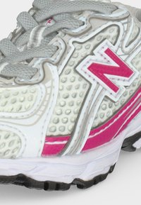 Gros plan sur une chaussure de sport blanche et rose avec un mesh texturé, des lacets gris et un logo rouge "N" bien visible sur le côté.