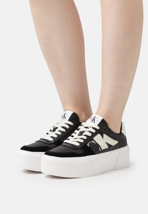 Sneaker piattaforme bianco e nero con una finitura lucida, caratterizzate da lacci a contrasto e un logo ben visibile su un lato.