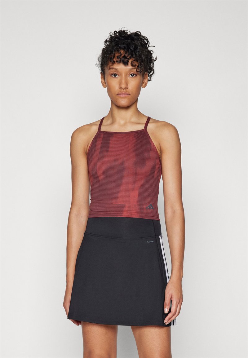Crop top de sport rouge avec une texture, associé à une jupe noire avec une fente latérale et un détail de rayure blanche.