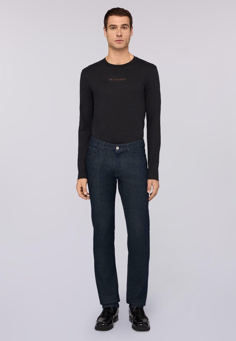 Camisa negra de manga larga con el logo "Trussardi"; pantalones de mezclilla azul oscuro con un corte clásico; zapatos negros pulidos; texturas suaves en todo el conjunto.