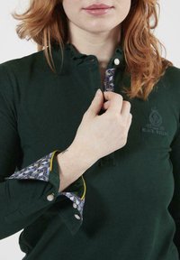 Polo vert avec un col à bouton, des poignets décorés de motifs avec des accents jaunes, un logo brodé et une texture douce.