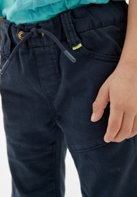 Námořnické modré kalhoty vyrobené z odolného materiálu s elastickým páskem na šňůrku, včetně předních kapes a bočních švů. Ruka spočívá na látce.