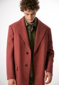 Czerwony wełniany płaszcz o klasycznym kołnierzu, zapinany na dwa guziki, z bocznymi kieszeniami, noszony na zielonej koszuli i prążkowanym golfie.