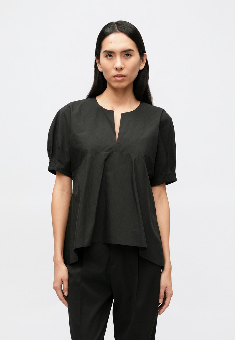 Hosszú, sötét hajú nő, laza fekete rövid ujjú, V-nyakú felsőben és fekete nadrágban, egyszínű világos háttér előtt állva.