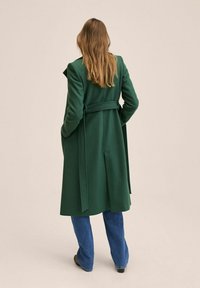 Lang grøn frakke med bælte, tekstureret stof, hakket krave og knælængde silhuet. Kombineret med lyseblå jeans og brune sko.