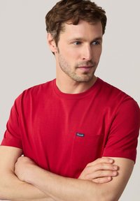 Rotes Baumwoll-T-Shirt mit kurzen Ärmeln, Rundhalsausschnitt und einer kleinen Brusttasche mit einem blauen Etikett. Glatte Textur, lässige Passform.