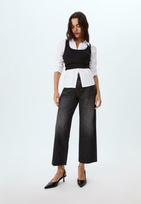 Top corto nero sopra una camicia bianca con bottoni, abbinato a jeans a gamba larga in denim scuro a vita alta e décolleté neri a punta.