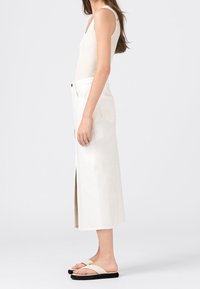 Bílá denimová maxi sukně s bočním rozparkem, v kombinaci s přiléhavým krémovým tílkem a minimalistickými béžovými sandály. Hladká textura, neformální design.