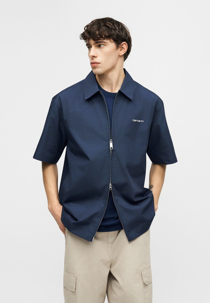 Jeune homme portant une chemise courte à manches courtes bleu marine zippée sur un t-shirt sombre et un pantalon cargo beige, les mains dans les poches, regardant vers la droite.