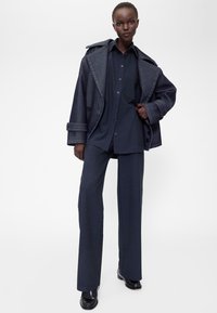 Sötét farmer anyagú, túlméretezett kabát nagyméretű hajtókával, hozzáillő sötét inggel és szabott nadrággal párosítva. Simogató tapintású, laza szabású.