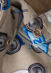 Col en tissu beige avec des poids de dessin animé imprimés, doté d'une doublure en polaire marron et d'une fermeture à bouton avec une surface texturée.