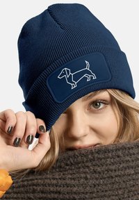 Navy knitted beanie with a smooth texture, featuring a rectangular patch displaying a white outline of a dachshund on the front.