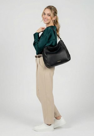 Femme portant un chemisier vert et un pantalon beige, avec un grand sac à bandoulière en cuir noir, souriante et debout devant un fond uni.