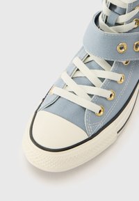 Blå kanvas høyhælte sneakers med hvite snører, gummitåhette og gull øyer. Har et bånd over ankelen for ekstra støtte.