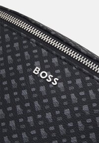 Černá texturovaná taška s kovovým zipem a logem "BOSS" v metalické barvě na vzorovaném povrchu.