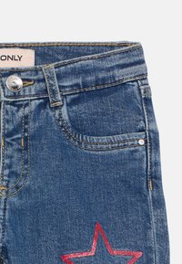 Jeans de ganga em azul com um bolso frontal, botão prateado e um detalhe em estrela de glitter vermelho na perna direita. Costura em contraste por toda a parte.