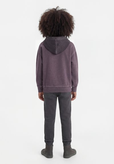 Bambino con capelli ricci che indossa una felpa con cappuccio viola sbiadita, pantaloni grigio scuro e scarpe da ginnastica alte grigie, in piedi di spalle su sfondo bianco.