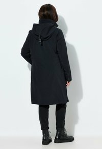 Personne portant un long manteau à capuche noir, un pantalon noir et des bottes noires à lacets, debout face à un mur blanc.