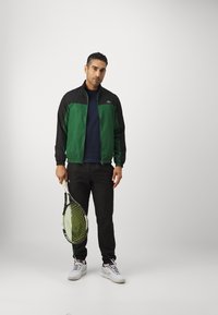 Jachetă verde și neagră cu fermoar peste un tricou bleumarin, asortată cu pantaloni negri și adidași albi. Ține o rachetă de tenis neagră cu șnururi galbene.
