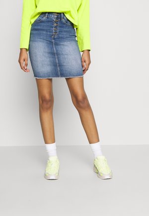 Personne portant un haut à manches longues vert néon, une jupe en jean taille haute boutonnée devant, des chaussettes blanches et des baskets jaune néon, debout sur un fond uni.