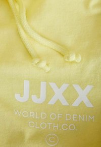 Un tejido amarillo con una textura suave presenta el texto impreso en blanco "JJXX WORLD OF DENIM CLOTH CO." y dos cordones a juego.