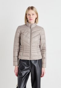 Lys beige, quiltet dunjakke med høy krage, frontglidelås og horisontale/chevron-sømmer. Båret med svarte bukser.