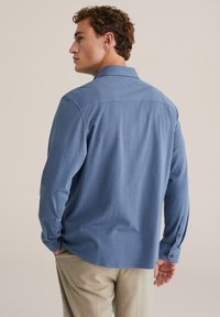 Világoskék ing gallérral, texturált anyagból, hosszú ujjú, kontrasztos barna gombokkal, bézs nadrággal párosítva.