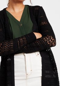 Black knit cardigan with openwork pattern and long sleeves, layered over a green blouse. White high-waisted pants visible at the bottom.