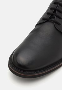 Chaussure de ville en cuir noir avec bout rond, lacets noirs et une subtile nuance brun rougeâtre sur le bord de la semelle.
