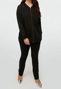 Giacca nera con zip frontale e maniche in pizzo a motivo, abbinata a pantaloni neri aderenti e décolleté argento con punta a punta. Tessuto morbido su tutta la superficie.