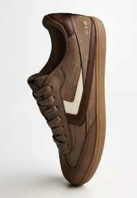Brown suede sneaker with dark brown leather accents, white side logo, lace-up front, and a soft rubber sole. Simple, streamlined design.