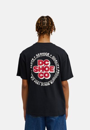 Personne avec des petites tresses porte un t-shirt noir avec un grand logo rouge et blanc "DC SHOE CO" et un texte circulaire dans le dos, assorti à un jean bleu.