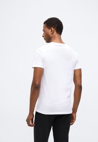 Rövid göndör hajjal rendelkező férfi, sima fehér, rövid ujjú pólóban és fekete nadrágban, egy fehér háttérnek háttal állva, a kamerának háttal.