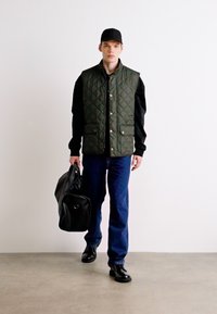 Gilet imbottito verde con bottoni dorati indossato sopra una felpa nera, abbinato a jeans blu e scarpe nere. Porta una borsa a tracolla nera.