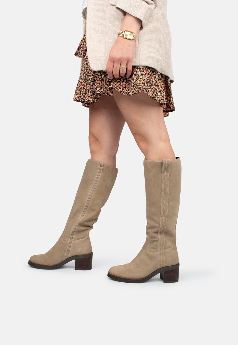 Botas de ante color beige hasta la rodilla con tacón bloque y cremallera lateral. El diseño incluye detalles de costura sutiles y una textura suave.