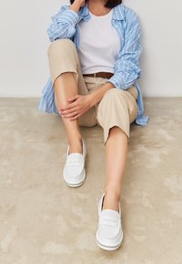 Scarpe bianche slip-on con dettagli perforati e suola testurizzata, abbinate a pantaloni beige chiaro e una camicia a righe blu.