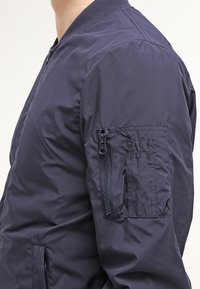 Tmavě modrá bomber bunda z hladké látky, s kapsou na zip na levém rukávu a žebrovaným límcem. Design je přiléhavý.