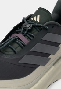 Μαύρο και σκούρο πράσινο αθλητικό παπούτσι με υφή στην επάνω επιφάνεια, ελαστικά κορδόνια και γκρι στοιχεία. Διαθέτει χαρακτηριστικό σχέδιο με τρεις ρίγες στο πλάι.