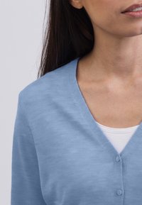 Kvinna som bär en ljusblå knappad kofta över en vit topp, visar sin hals och en del av sitt ansikte med långt mörkt hår.