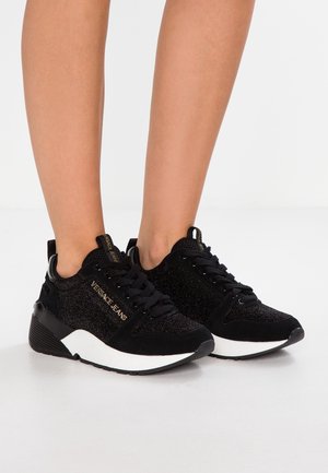 Jambes portant des baskets Versace Jeans noires pailletées avec semelles blanches sur un sol blanc, lacées et placées côte à côte.