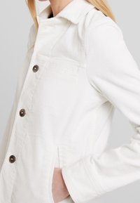White corduroy jacket featuring a collar, five buttons, two side pockets, and ribbed texture. The sleeve is cuffed at the wrist.