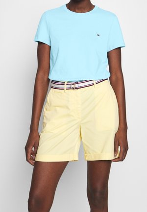 Osoba o ciemnej karnacji, ubrana w jasnoniebieską koszulę z krótkim rękawem włożoną do żółtych szortów z paskiem w paski, stojąca na jednolitym tle.