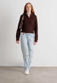 Bruine geribde trui met een hoge kraag, gecombineerd met lichtblauwe ripped jeans en witte schoenen. Een bruine schoudertas wordt over het lichaam gedragen.