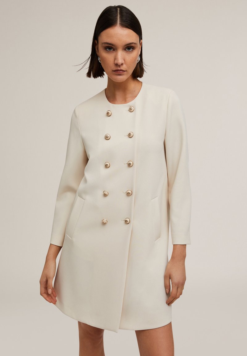 Rochie-closet beige, cu guler rotund, mâneci lungi, deschidere dublă în față și șase nasturi aurii, având buzunare laterale.