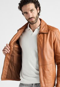 Brun læderjakke med en glat tekstur, der har en opretstående krave, lynlås lukning og indvendig lomme, båret over en hvid polo shirt.