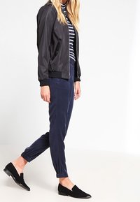Chaqueta bomber negra con puños acanalados, combinada con pantalones tapered azul marino y zapatos planos negros. Suéter de cuello alto a rayas debajo.