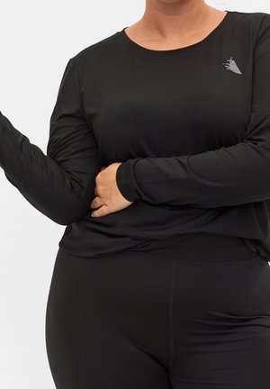 Person wearing black long-sleeve athletic top and black leggings, posing with one hand holding opposite arm across torso.