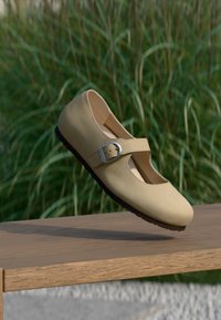 Μπεζ παπούτσι Mary Jane με λουράκι και τοκά, αιωρούμενο πάνω από ένα ξύλινο παγκάκι, φόντο ψηλό πράσινο γρασίδι.