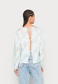 Blusa floral azul clara com mangas longas, costas abertas e laços, feita de um tecido texturizado. Combinada com jeans de denim azul claro.