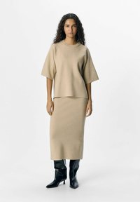Geweven beige top met korte mouwen, gecombineerd met een bijpassende rok tot halverwege de kuit. Het model draagt glanzende zwarte laarzen met puntigeneuzen. Soepele textuur.